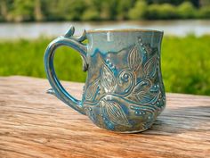 a blue coffee mug sitting on top of a wooden table next to a river and grass