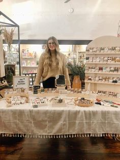 a woman standing in front of a table with many items on it and shelves behind her