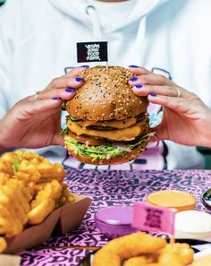 a woman holding up a hamburger and french fries at a table with other food items