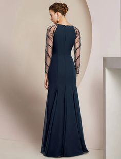 a woman in a long blue dress with sheer sleeves and an open back, standing against a