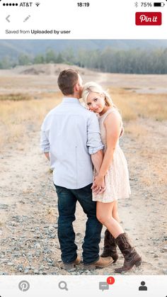 a man and woman standing in the middle of a dirt road with mountains in the background