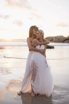 two women in white dresses hugging on the beach at sunset, with one woman standing next to her