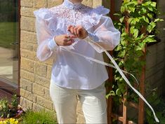 a woman standing in front of a brick building wearing white pants and a shirt with ruffles on it