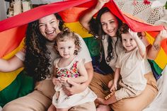 three women and two children sitting on a bed under a rainbow colored cloth over their heads