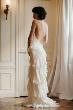 the back of a woman in a white wedding dress standing on a wooden floor next to a window