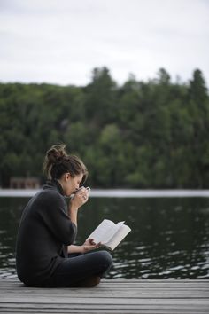 a woman sitting on the edge of a lake reading a book and holding a cup