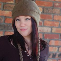 a woman with long hair wearing a beanie and looking at the camera while standing in front of a brick wall