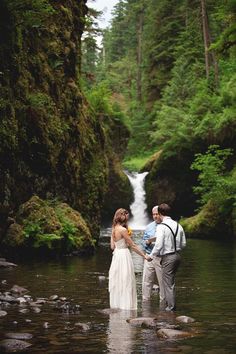 a bride and groom are standing in the water near a waterfall holding each other's hands