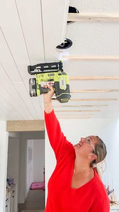 a woman in red shirt holding up a driller on the ceiling above her head