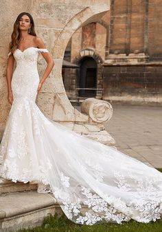 a woman in a white wedding dress standing next to a stone wall with her hands on her hips