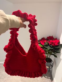 a hand holding up a red crocheted purse with flowers in the corner behind it