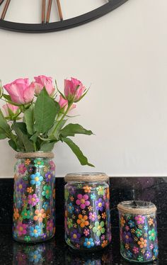 three jars with flowers in them sitting on a black counter top next to a clock