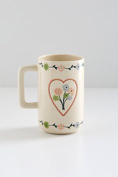 a white coffee mug with flowers painted on the side and a heart - shaped handle