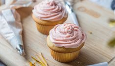 two cupcakes with pink frosting sitting on a cutting board