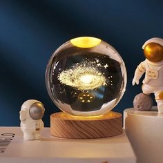 an astronaut figurine standing next to a glass ball with fireworks in it on a wooden base