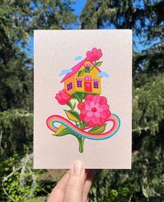 a hand holding up a card with a house and flowers in the center, surrounded by trees