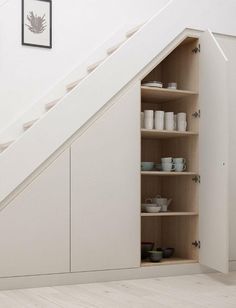 an open cabinet under the stairs in a room with white walls and wooden floors, filled with dishes