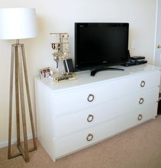 a flat screen tv sitting on top of a white dresser next to a floor lamp