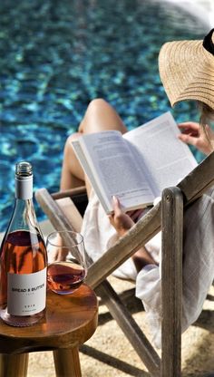 a woman sitting in a chair next to a pool reading a book and drinking wine