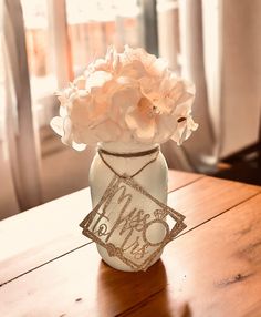 a white vase with some flowers in it on a wooden table next to a window