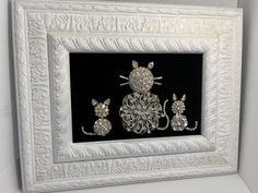 an ornate white frame with three pieces of jewelry in the shape of cats and mouses