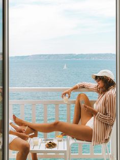 a woman sitting on top of a white chair next to a window near the ocean