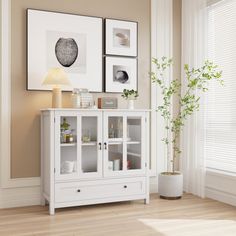 a white cabinet with glass doors next to a plant and pictures on the wall above it