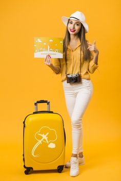 a woman in white pants and a hat holding a sign next to a yellow suitcase