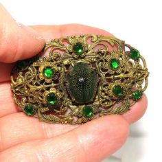 An early 1900's filigree brass pin with a flat C catch. The filigree is set with green rhinestones and has embossing on the back too. In the center is a real green leaf beetle. The pin is in excellent condition and measures 5.3 by 3.4 cm. Ornate Oval Filigree Brooches, Ornate Bronze Brooch Jewelry, Green Art Nouveau Wedding Brooches, Vintage Bronze Brass Brooches, Antique Green Jeweled Jewelry, Victorian Green Collectible Brooches, Antique Jeweled Brooch For Wedding, Antique Gold Brass Brooches, Vintage Bronze Jeweled Jewelry