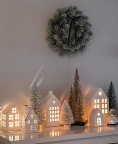 lighted christmas trees and small houses on a shelf in front of a wall mounted clock