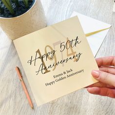 a hand holding a happy 50th anniversary card next to a potted plant and envelope