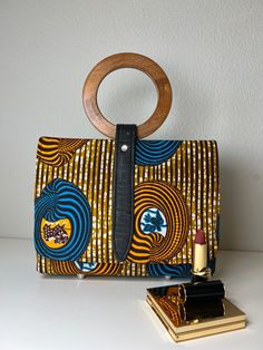 This gorgeous Aisha African Print Hand Bag is perfect for travel, work, or parties. Our light bag is sourced from Ghana and made of Authentic African print fabric with stitch details for a luxe look and are wooden handle. The Interior accessory and pockets help you keep items organized in your purse. This bag is individually handcrafted and lovingly made to be unique to you Description: African Print handbag with Wooden handle The inner is fully lined with black fabric Interior compartments for Luxury Bags With Bamboo Handle For On-the-go, On-the-go Clutch Box Bag With Detachable Handle, Modern Travel Box Bag With Round Handle, Travel Box Bag With Top Leather Handles, Rectangular Shoulder Bag With Bamboo Handle For On-the-go, On-the-go Shoulder Bag Satchel With Bamboo Handle, Daily Use Satchel With Round Bamboo Handle, Everyday Box Bag With Round Handle, Bamboo Handle Shoulder Satchel