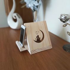 a wooden phone holder with a cat sitting on it's side, next to a flower vase