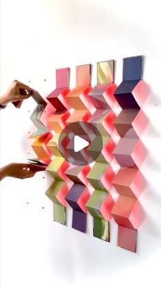 two hands reaching for an art piece made out of colored papers and paper strips on a white wall