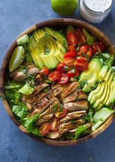 a salad with chicken, avocado and tomatoes in a wooden bowl next to a glass of dressing