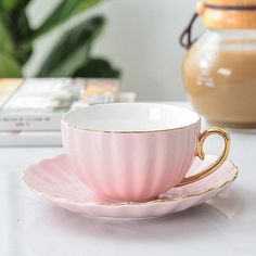 a pink tea cup and saucer on a white table with a potted plant in the background