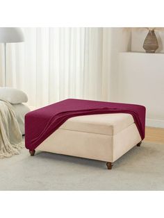 a white ottoman with a red blanket on it in front of a bed and lamp