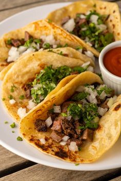 three tacos on a white plate with sauce and garnishes next to it