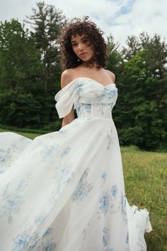 a woman in a white and blue dress is standing on grass with trees behind her