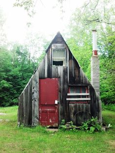 an old wooden building with a red door and window in the grass next to trees