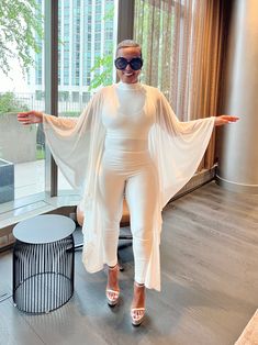 This elegant ensemble combines a sleek, form-fitting white jumpsuit with a flowing, sheer cape, creating a striking blend of modern sophistication and timeless grace. The outfit's minimalist design is accentuated by its clean lines and pure white fabric, making it perfect for high-profile events or stylish gatherings. The sheer cape adds a touch of ethereal charm, while the overall silhouette flatters the figure, offering both comfort and style. Paired with bold accessories, this outfit exudes c White Long Sleeve Jumpsuits For Night Out, White Long Sleeve Jumpsuits For Evening, White Long Sleeve Jumpsuits And Rompers For Evening, Sheer Cape, Cape Jumpsuit, Outfits Minimalist, Plus Jumpsuit, Bold Accessories, Fabric Making