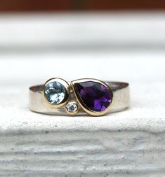 This is a multistone birthstone ring. This particular ring features an amethyst, aquamarine and diamond, but it can be made with other combinations of stones and the shapes could also be changed.If you get in touch we can work out options and prices for you. The ring is recycled sterling silver in a 4mm wide band. The pear shaped amethyst is 5x7mm and the aquamarine is 4mm round with a 1.5mm diamond, all set in recycled 9ct yellow gold.The ring is made to order in the size you need. You can leav Modern Ring Design Women, Family Birthstone Ring Ideas, Aquamarine And Amethyst Ring, Modern Multi-stone Birthstone Ring For Anniversary, Modern Topaz Ring With Gemstone Accents For Anniversary, Amethyst Birthstone Ring With Bezel Setting, Modern Diamond Amethyst Ring, Purple Multi-stone Diamond Ring, Modern Amethyst Diamond Ring