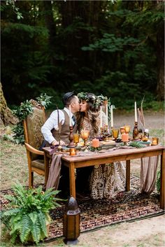 a bride and groom kissing at the end of their dinner table surrounded by greenery