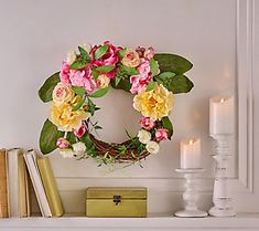 a wreath with pink and yellow flowers on top of a mantle next to two candles