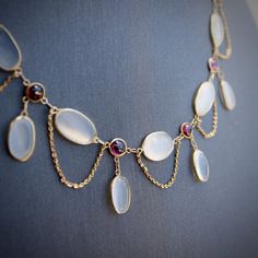 This is part of Chairish’s Fine Jewelry assortment.  This gorgeous festoon necklace has a soft and delicate arrangement of moonstone and garnet cabochons, favourites of the Edwardian era. Feminine and romantic, the necklace curtains to create gorgeous curved lines around the neck. Five oval moonstone cabochons lay horizontally across a hand-made 14k gold chain, while four oval drop shaped moonstones dangle gracefully from alternating garnets. The gold chain loops between the cabochons for a lace Edwardian Era Jewelry, Jewelry With Gemstones, Moonstone Beaded Necklace, Art Nouveau Necklace, Edwardian Jewelry Necklaces, Antique Amethyst Jewelry, Moonstone Necklace Vintage, 16th Century Jewelry, Moonstone Necklaces