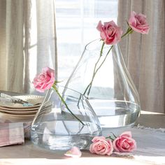 three pink roses in a clear vase on a table next to plates and napkins