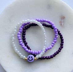 Purple beaded bracelet stack including 3 stretch bracelets: one light purple bracelet, one dark purple bracelet, and one pearly white bracelet with a purple smiley face surrounded by galvanized gold seed beads. Please refer to the size chart to find your bracelet size. Purple And White Beaded Bracelet, Cheap Purple Bracelets With Tiny Beads, Light Purple Bracelet, Purple And White Bracelet, Trendy Purple Beaded Bracelets For Everyday, Everyday Purple Beaded Stretch Bracelet, Trendy Everyday Purple Beaded Bracelets, Purple Round Beads Friendship Bracelets, Purple Stretch Bracelet With Round Beads For Everyday