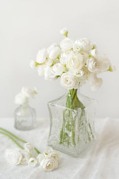 white flowers are in a clear vase on a lace tablecloth with two small glass vases