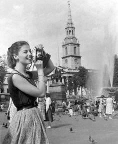 an old photo of a woman holding a camera in front of a fountain with pigeons around her