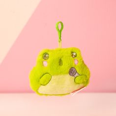 a small green purse hanging from a hook on a pink and white wall with a clip in the shape of a frog